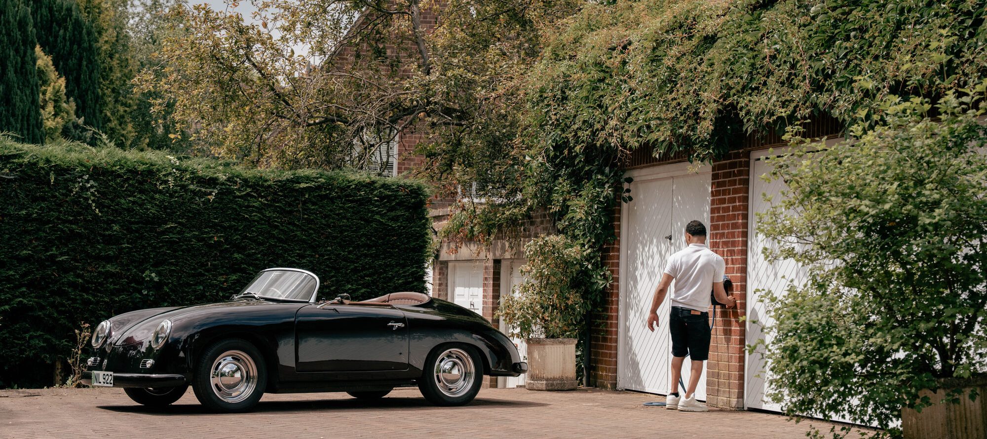 A classic black convertible parked on a brick driveway while its owner unhooks a charging cable beside the garage